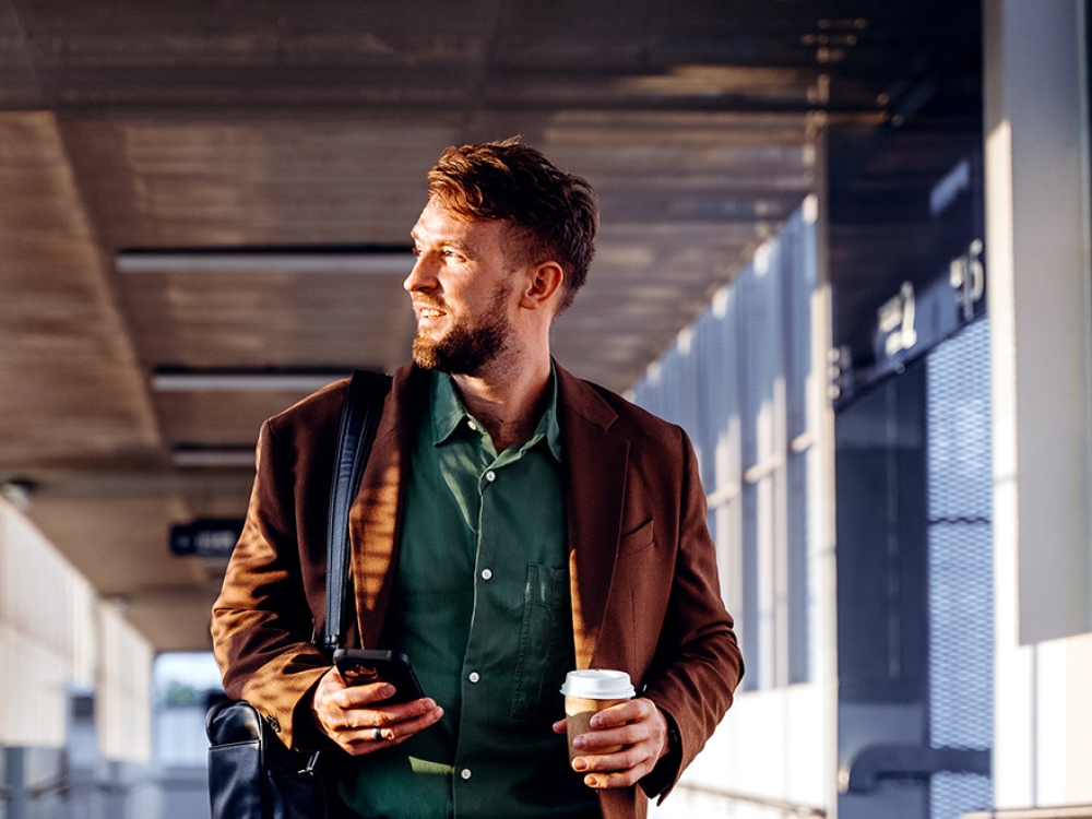 A person walks through a covered corridor, holding a smartphone and a coffee cup, with a black bag over their shoulder.